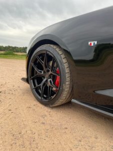 Close-up of a black sports car's wheel and tire with red brake calipers on a dirt road.