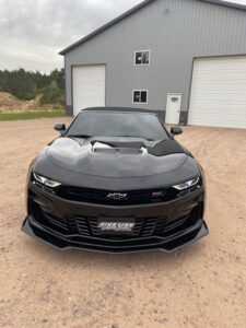 A sleek black sports car parked on a dirt driveway in front of a gray building.